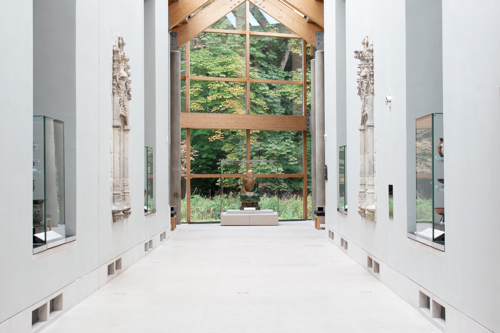 A statue on display at the end of a spacious gallery at the Burrell Collection in Glasgow. Trees can be seen through the glass walls at the back of the gallery.