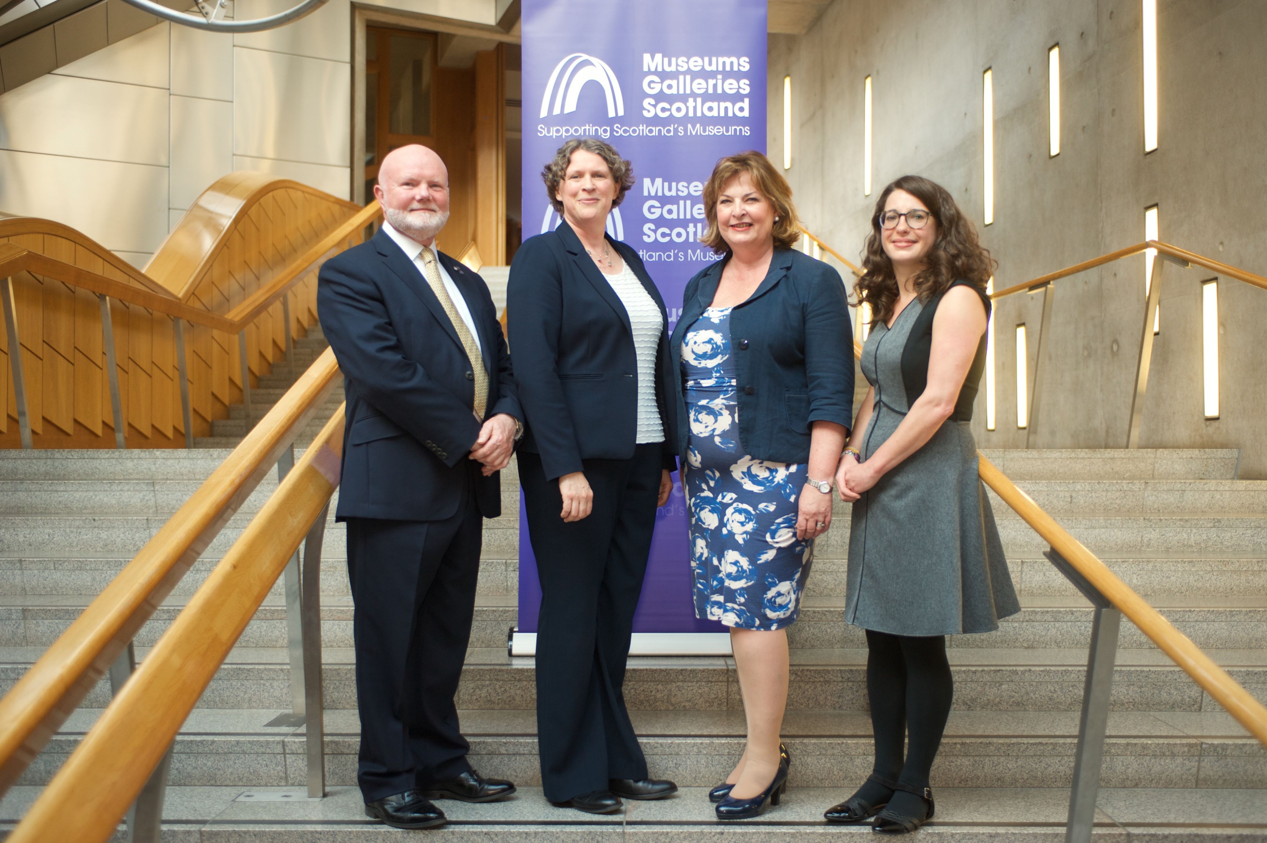 Four people in smart outfits standing in front of a Museums Galleries Scotland banner on concrete steps in the Scottish Parliament.