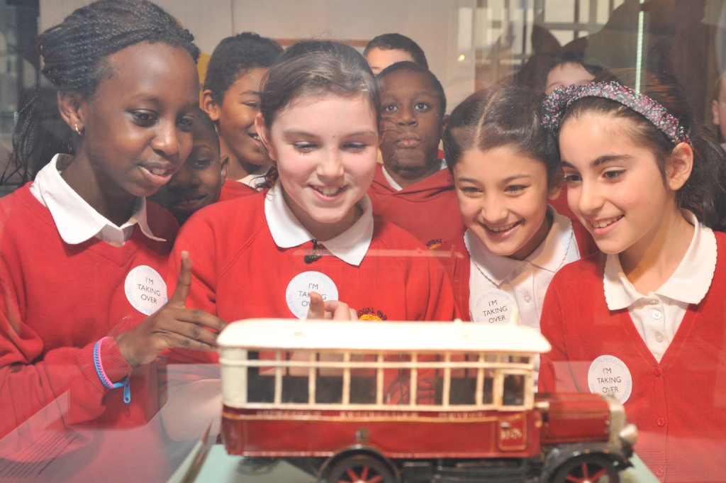 A group of children in red school uniforms pointing at a model bus on display in a museum case.