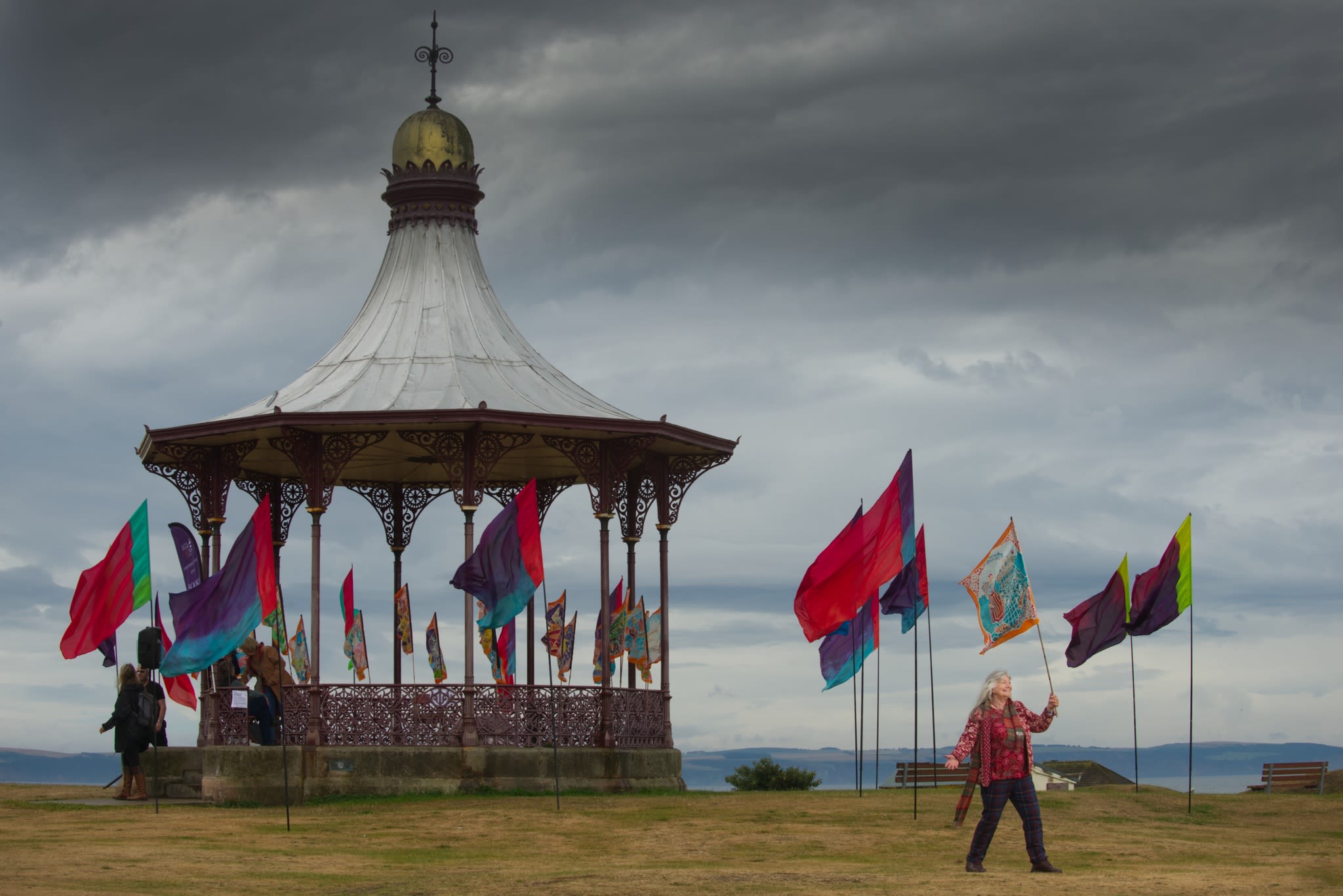 A woman waves a flag in front of a bandstand. The bandstand is surrounded by colourful flags on poles. Image credit: VisitScotland/Rob McDougall.