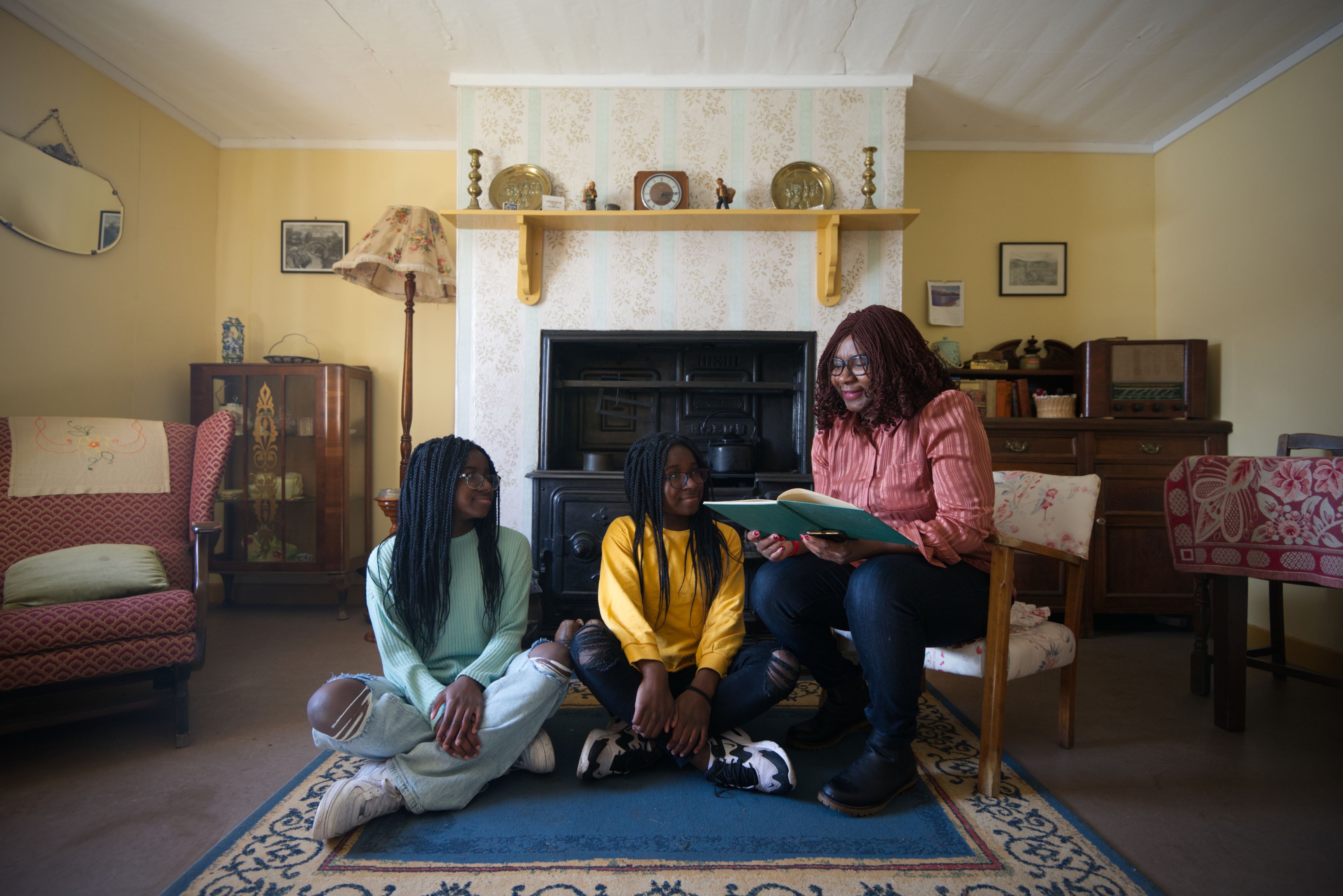 A seated woman reads a book to two teenage girls in a 20th century living space at the Highland Folk Museum.